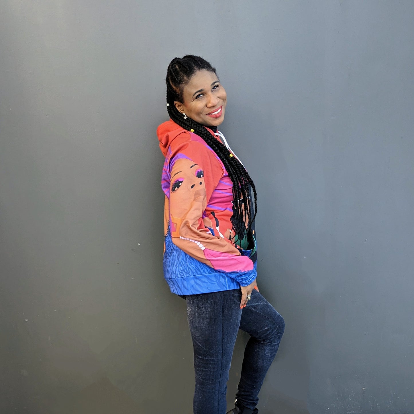 Lachi, a black woman with cornrows, poses wearing a colorful sweater featuring the album art for her single Black Girl Cornrows which has drawn profiles of Lachi and the other artists over an orange background
