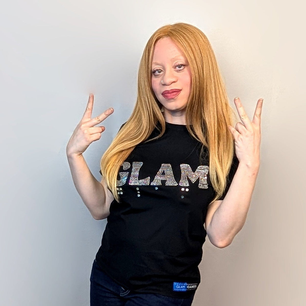 An albino woman poses wearing a shirt that reads "GLAM" in rhinestones with rhinestone braille under the word