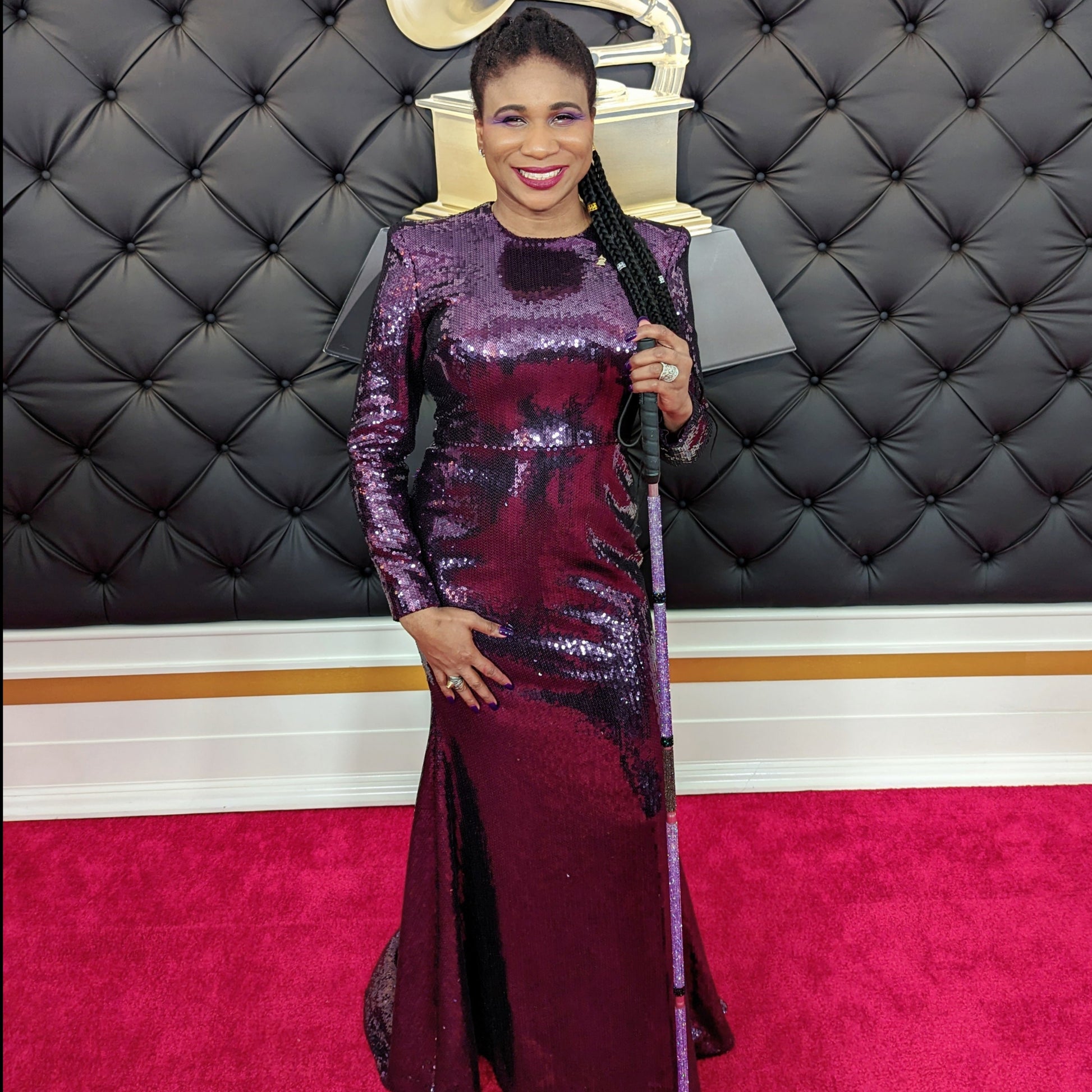Lachi, a black woman with cornrows, poses at the Grammy Red Carpet in an elegant purple dress and purple rhinestone glam cane
