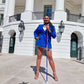 Lachi, a black woman with cornrows, poses confidently in front of the White House Rotunda, wearing a blue suit jacket and a white skirt, holding a red white and blue rhinestone glam cane