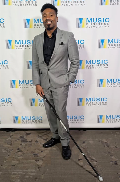 Deshaymond, a handsome black man in a gray suit holding a black rhinetone glam cane on a red carpet 